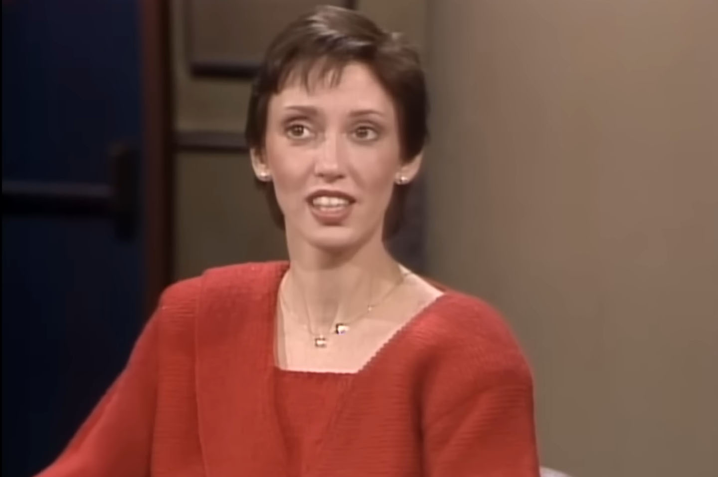 Close-up of actress Shelley Duvall speaking during an interview, wearing a red sweater, with a thoughtful expression